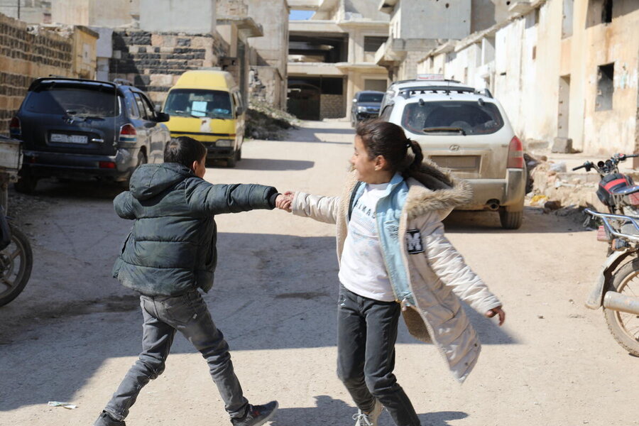 Dos niños juegan, cogidos de la mano, en una calle en la que se ven vehículos aparcados y edificios. El chico lleva un anorak negro y la chica, un abrigo blanco.